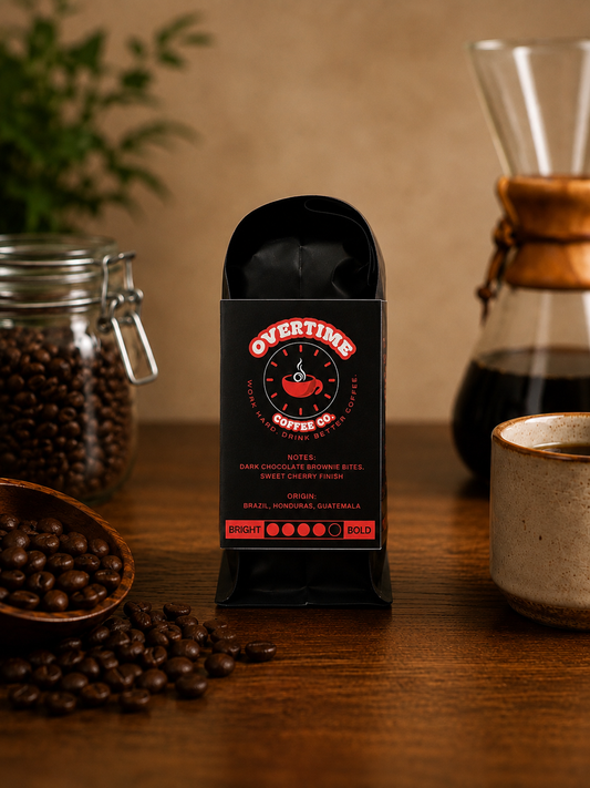 A bag of Overtime Coffee Co. dark chocolate brownie bite coffee beans stands on a wooden table, surrounded by coffee beans, a glass carafe, and a ceramic cup in a warm, rustic setting.