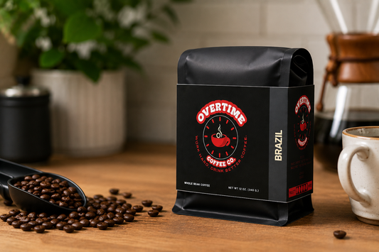 A black bag of Overtime Coffee Co. whole bean coffee stands on a wooden table, surrounded by scattered coffee beans, a coffee scoop, and a coffee maker in a warm kitchen setting.