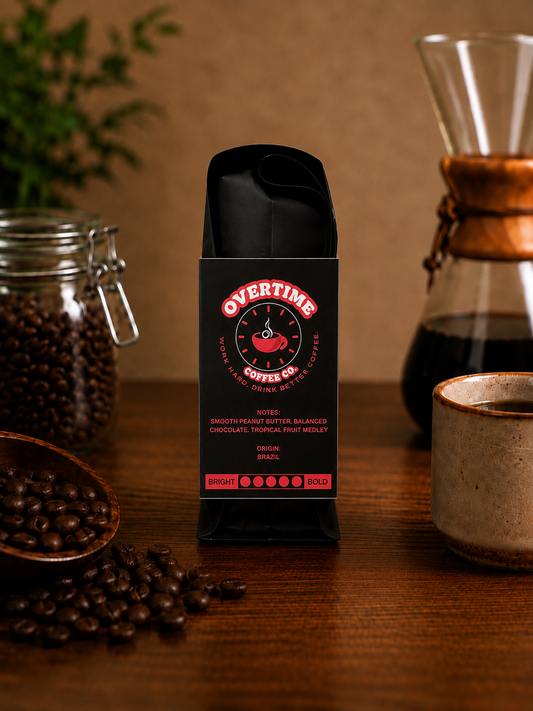 A bag of Overtime Coffee Co. coffee stands on a wooden table, surrounded by coffee beans, a glass carafe, and a ceramic cup in a warm, rustic setting.